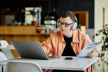 A man with down syndrome works on his laptop in a cafe, headphones on, focused on his work.