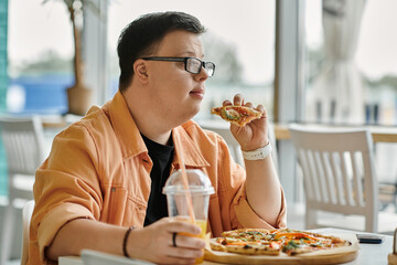 A man with down syndrome sits at a cafe table, taking a bite of a delicious pizza slice.