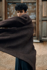 Fashionable young man in brown coat and scarf standing in front of window with arms outstretched