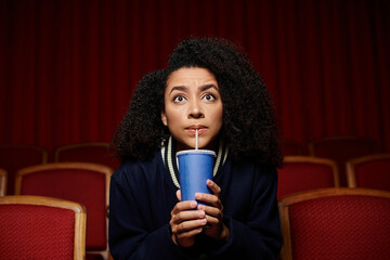 A young woman in casual clothes sits in a theater seat, holding a cup and staring up in surprise,...