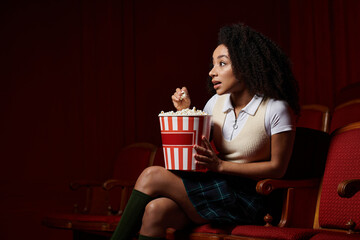 A young woman captivated by the film on screen, sits in a theater seat with a bucket of popcorn,...