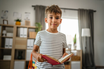 Portrait of young boy stand and hold, read book in the living room