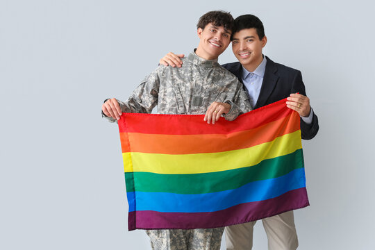Young soldier with his husband and LGBT flag on light background