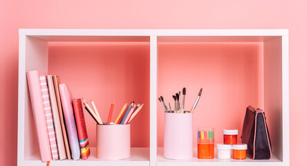 School supplies and books stand on a white shelf against a pink background.