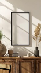 An empty frame with a black border stands on a wooden cabinet, surrounded by pottery and a cup, bathed in sunlight and shadows, creating a calm, rustic ambiance.