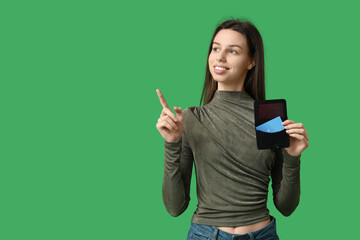Thoughtful young woman holding credit card holder and pointing at something on green background