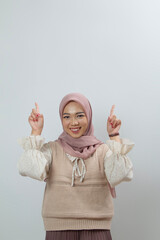 young woman in hijab in light beige knitted vest top posing with both hands pointing upwards in a white isolated room