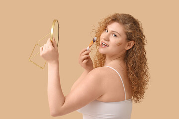 Beautiful young woman with foundation on her face holding mirror and makeup brush against beige background