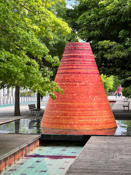 The iconic red and orange tiled Volcano Fountain next to a water channel and park benches along the Alameda dos Oceanos promenade in Parque das Nacoes, Lisbon, Portugal.