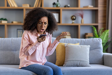 Woman sitting on couch expressing frustration while looking at smartphone screen in modern living...