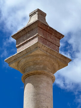 A low-angle close-up shot of the capital and top decorative element of the historic stone pillory in Alhandra, Portugal, set against a bright blue sky with white clouds.