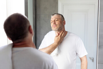 Obraz premium Ingrown hair problem. Unshaved middle-aged man touching neck, looking at mirror in bathroom, making morning beauty routine