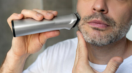 Middle aged man trimming beard with electric trimmer indoors in bathroom, closeup cropped shot