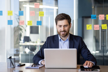 Fototapeta premium Successful smiling man working with laptop inside office, satisfied with results businessman typing on keyboard, mature boss in business suit at workplace.