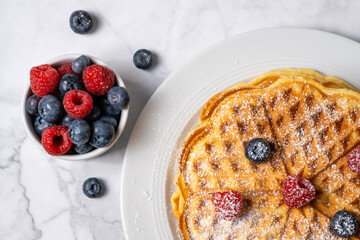 Top view of Belgian waffle and mix of raspberries and blueberries on marble background. Delicious summer breakfast. Stack of homemade round waffles and berries, flat lay