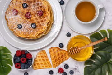 Belgian waffles, berries, honey and cup of tea: delicious summer breakfast from above. Heart-shaped waffle and mix of raspberries and blueberries as topping on marble background. Flat lay, top view