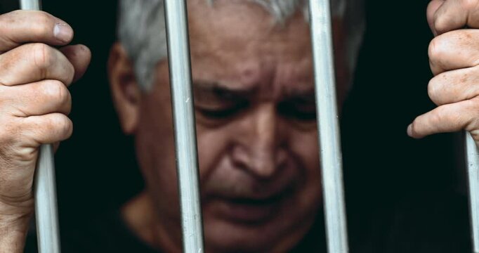 Imprisoned senior gray hair man crying in despair behind metal bars feeling desperate in old age entrapped by sorrow and past regrets, close-up of hand gripping metal bar