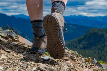 A closeup look at male hiking boots, hiking on top of a mountain on a sunny day with rugged mountains in the background in the Pacific Northwest.