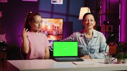 Daughter and mother in home studio film video together for social media channels, holding green screen laptop on desk promoting sponsor. Mom and her young kid recording content next to mockup device