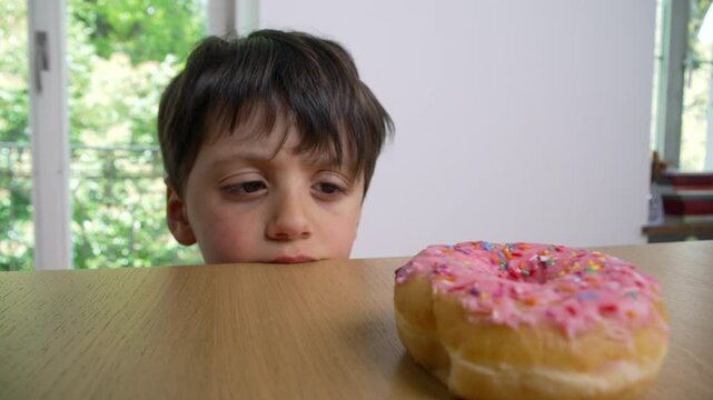 Boy stares at a pink frosted donut with colorful sprinkles on a wooden table, representing childhood desires and indulgence, focused and longing indoors