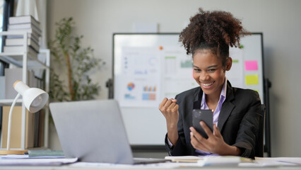 Young professional woman celebrates success in a modern office, fully engrossed in her smartphone, exuding happiness and excitement as she embodies the essence of a successful businesswoman
