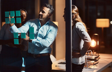 Night, brainstorming and business people with glass board for discussion, project ideas or sticky note in office. Writing, diversity and employees in meeting for planning, agenda or collaboration