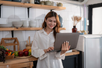 Young woman is using a laptop while standing in a kitchen with fresh food on the counter behind her. She is smiling and appears to be enjoying her work