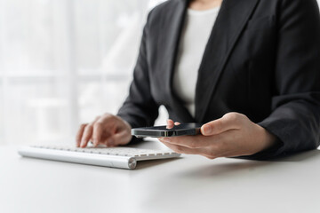Businesswoman multitasks in her office, holding a smartphone while typing on a wireless keyboard, showcasing efficiency