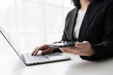 Businesswoman is multitasking, holding a smartphone in one hand and typing on a laptop keyboard with the other, showcasing efficiency and connectivity in the modern workplace