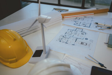 Scale model of a wind turbine is standing on an architect's desk amidst blueprints and drafting...