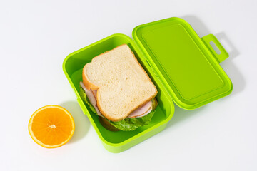 Green lunch box with ham sandwich inside and an orange on white background