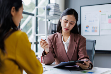Fototapeta premium Two cheerful asian office workers women cooperating on project, sitting at desk with laptop using calculator, finance accounting analytical concept.