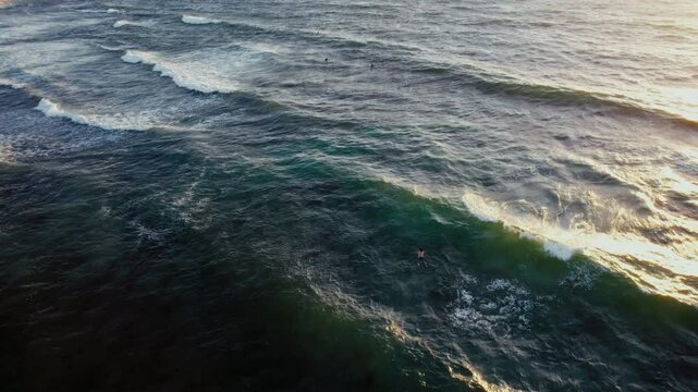 A lone figure paddles through the water as waves crash along the coastline. The sun shines brightly on the water, creating a glistening effect.