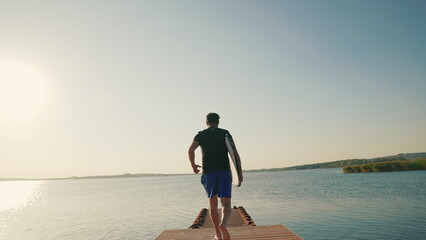 A male surfer runs along a pier on a lake against the sun, holding a water board. A male surfer walks along the pier with a small surfboard in his hands. Summer water sports