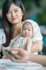 Obraz premium A twenty-something Malaysian woman holding a few-months-old baby at a stylish cafe in Selangor, Malaysia, on a sunny early summer afternoon.
