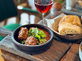 Close up of a rustic wooden table with black bowl of braised meat in tomato sauce garnished with fresh cilantro, accompanied by crusty bread and glass of red wine, set in cozy restaurant ambiance.