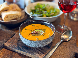 Close up of a rustic wooden table with a bowl of creamy pumpkin soup garnished with pumpkin seeds and a microgreen leaf, served alongside crusty bread, a mixed green salad, and glass of red wine.