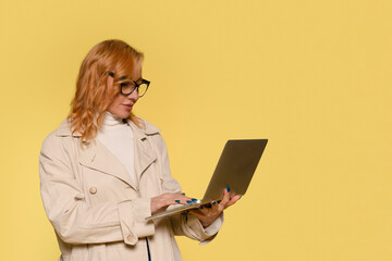 Woman in Beige Trench Coat Uses Laptop in Front of Yellow Background with Copy Space