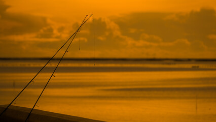 Serene sunset over calm waters with fishing rods silhouetted in the foreground, capturing a peaceful evening by the coast.