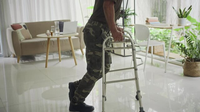 Tilt up shot of male veteran in military uniform using walker during rehabilitation at home after war