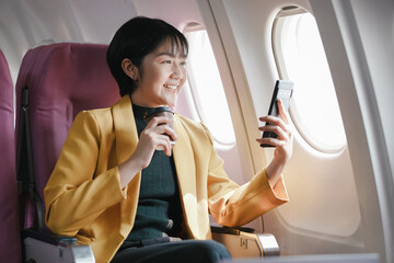 A woman is sitting on an airplane, smiling and holding a cell phone. She is also holding a cup of coffee