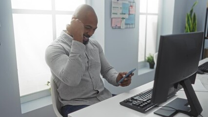 Young man in office checking smartphone with a smile on his face while sitting at a desk with a computer behind him