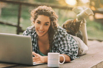 woman with curly hair in a braid, dressed in a checkered shirt, lies on a bench using a laptop. A cat sleeps on her back, and a coffee mug is nearby, emphasizing a cozy outdoor work setting