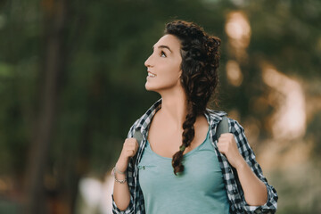 Young woman with curly hair in a braid, dressed in a checkered shirt and green top, smiles while looking around. Her backpack indicates a casual and adventurous outdoor activity