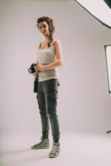 Confident woman with curly hair in a braid, wearing a sleeveless top and cargo pants, holds a camera. She stands in a studio, exuding a strong sense of pride and enthusiasm for her photography role