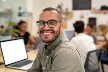 Fototapeta premium A man in a green shirt and glasses smiles confidently while working on his laptop in a modern office.
