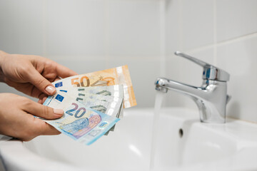 Women's hands hold money against the background of a sink and a faucet with water. Concept of utilities, payment and savings.