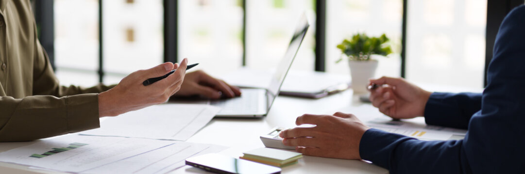  professional setting featuring two individuals working at a table. They are engaged in reviewing documents and using a calculator. Laptops are open on the table.