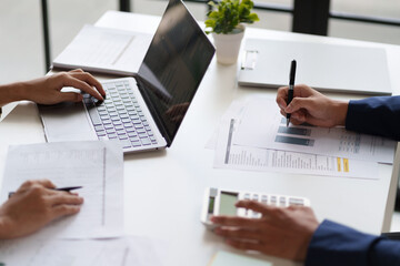  professional setting featuring two individuals working at a table. They are engaged in reviewing documents and using a calculator. Laptops are open on the table.