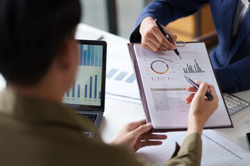 Two individuals are engaged in a business discussion, focusing on a document with graphs and charts. One person is pointing with a pen while the other is holding a clipboard